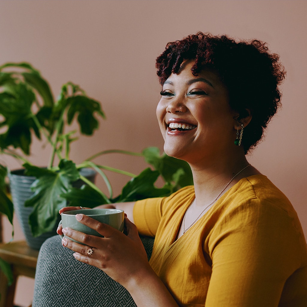 Photo of a lady with a nose piercing, sitting on a sofa by a plant holding a mug