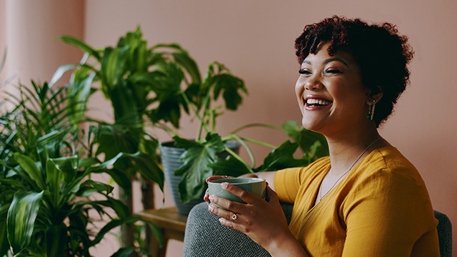 Photo of a lady with a nose piercing, sitting on a sofa by a plant holding a mug